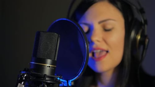 A Young Woman in Earphones Sings a Song Near a Microphone in a Recording Studio Under Natural Light
