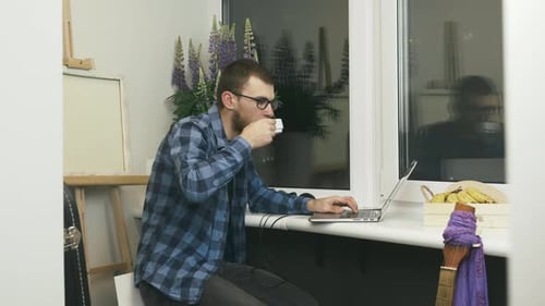 Junger Geschäftsmann benutzt Laptop am entfernten Heimarbeitsplatz auf dem Balkon und trinkt Kaffee