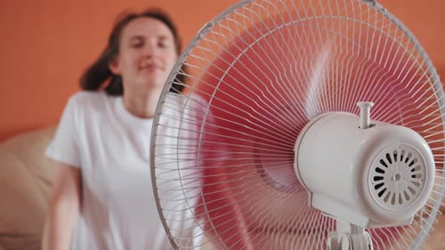 Young Woman Sits in Front of Electric Home Fan Air Stream Blowing Short Hair