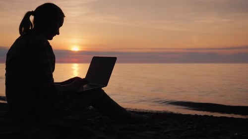 Side View of Silhouette of a Woman Working with a Laptop By the Sea at Sunset