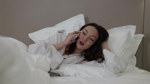 A Cheerful Brunette Woman in a White Shirt Is Lying in the Room on a Bed on White Bed Linen