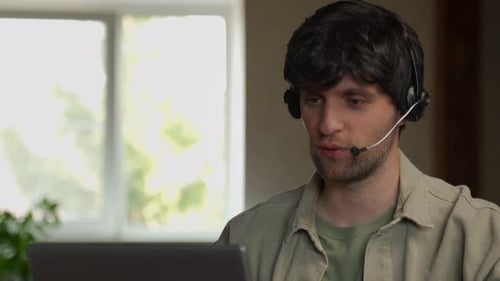 Joyful Young Adult Male Manager or Call Center Employee in a Headset Looks at a Laptop Screen While