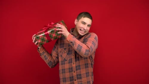 A Young Man Shakes a Christmas Present in His Hands and Rejoices. Shooting in the Studio on a Red