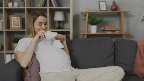 Pregnant Woman Smiling, Holding Baby Shoes on Couch