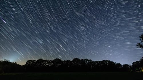 moving night sky with star trails over rural landscape with field in front of trees