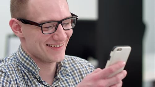 Young Man in Office Laughing at Text Message on Smartphone