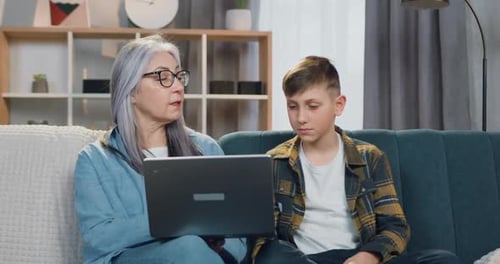 Abuela mayor con gafas sentada junto con su nieto y él con los deberes en la computadora