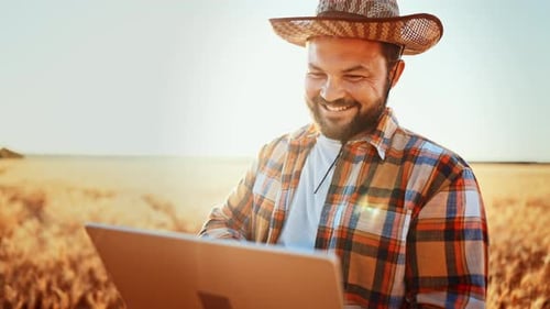 Caucasian Farmer with Beard in Straw Hat Standing in Wheat Field and Typing on Keyboard of Laptop