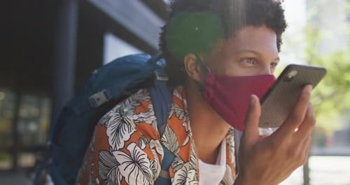 Hombre afroamericano en la ciudad, con máscara facial sentado con un teléfono inteligente en la calle