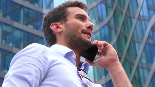 A Young Handsome Businessman Talks on a Smartphone - Closeup From Below - an Office Building