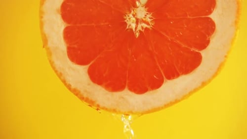 Water flows down from a slice of grapefruit on an orange background. Slow motion.