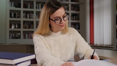 Ein studentisches Mädchen mit Brille sitzt mit Büchern und Schreiben an einem Bibliothekstisch