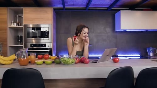 Woman Using Laptop and Eating Apple in Kitchen