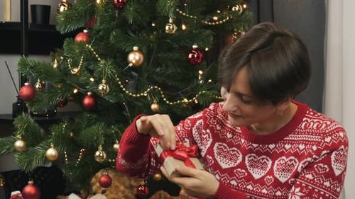 Woman Opening Christmas Gift by Decorated Christmas Tree