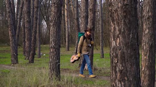 A Backpacker with a Guitar and a Camera Walks Along a Trail in the Forest