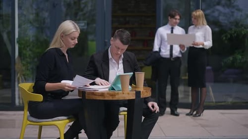 Concentrated Young Coworkers Sitting on Terrace Analyzing Project As Blurred Colleagues Standing at