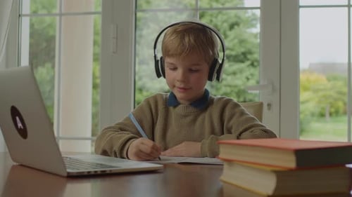 School Boy Writing Looking at Laptop Sitting at the Table By the Window