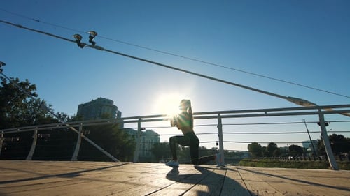 Active woman silhouette exercising and stretching in city park on footbridge.