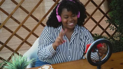 Young African woman having video call with mobile smartphone cam while sitting in bar
