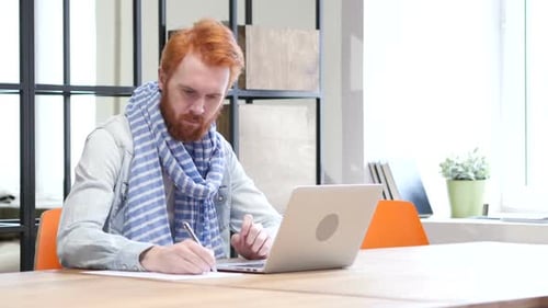 Man With Red Hair Working at Desk With Laptop