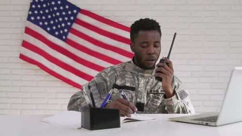 Soldier Writes and Speaks into Walkie-Talkie by Flag