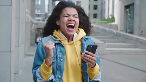 Excited woman celebrates success using smartphone outdoors