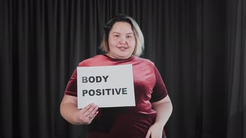 Eine selbstbewusste, flirtende, übergewichtige Frau hält ein Typenschild mit einem Schild KÖRPER POSITIV