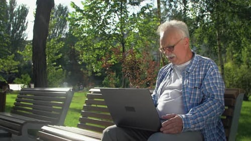Angry Senior Man Talks in Skype, Sitting on the Bench in the Garden. A Pensioner Is Holding a Laptop