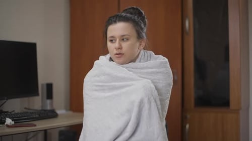 Jeune femme malade enveloppée dans une couverture frissonnant regardant autour de elle à l'intérieur de la maison