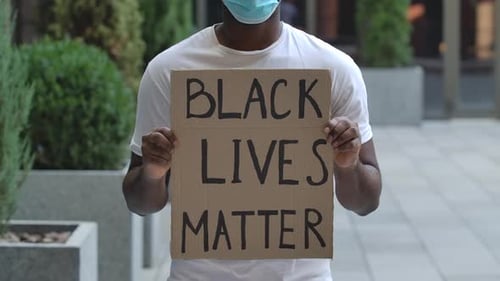 An African American Stands in the Middle of the Street Holding a Cardboard Poster in Front of Him