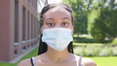 A Young Black Woman in a Face Mask Looks at the Camera - Face Closeup - an Office Building