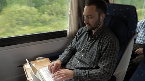 Junger Mann arbeitet hinter einem Laptop und sitzt in einem Zug.