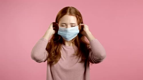 Young Pretty Woman Puts on Face Medical Mask During Coronavirus Pandemic. Portrait on Pink