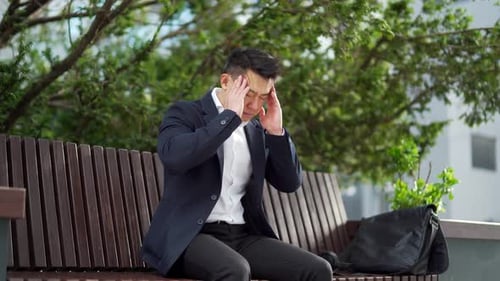 exhausted young asian business man office worker sitting on bench in city park outdoors.