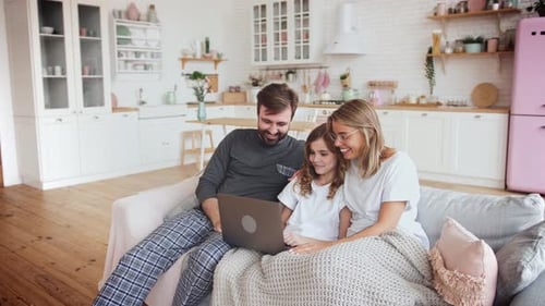 Happy Family Sitting on Grey Sofa and Using Laptop in Big Living Room with Kitchen on Background