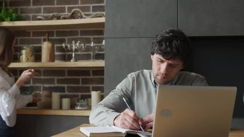 Woman Approaches Man Working at Laptop in Kitchen