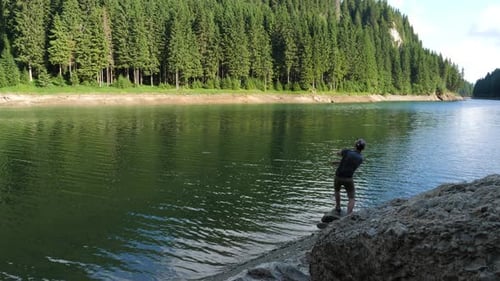 Young man throws the stone in the water of a mountain lake in the forest.