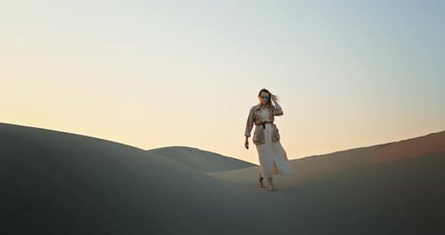 Attractive Girl in Long Dress Walking Down the Sand Dunes in Desert at Sunset