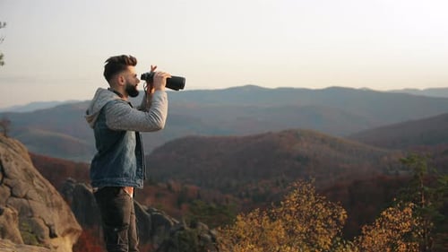 The Man is Standing on the Edge of a Cliff and Looks Through Binoculars