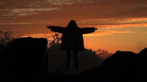 Woman Silhouette with Arms Outstretched on Rocky Beach at Sunset