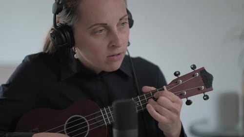Close up of Serious woman in a blue shirt and headphones records a lesson on playing ukulele