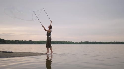 A Young Female Artist Shows a Soap Bubble Show Blowing Up Huge Soap Bubbles on the Shore of a Lake