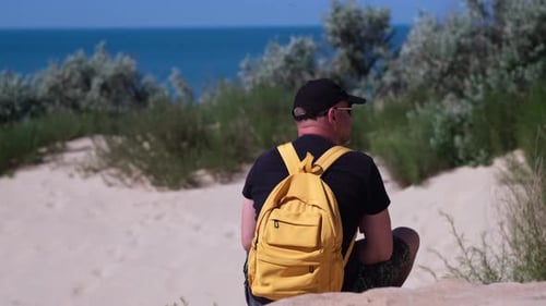 Man Traveler with Backpack Sits on Sand Beach Dune Looking at Amazing Sea View