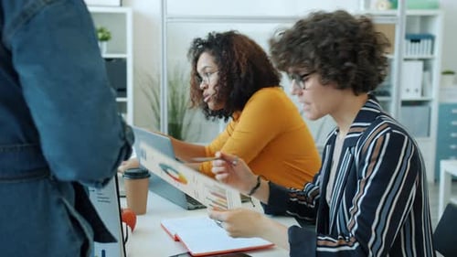 Cheerful Women Discussing Work Looking at Papers and Sharing Ideas Sitting at Desk in Office