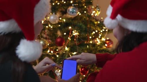Two Females in Green Santa Hat Using Smartphone on Christmas Tree Background