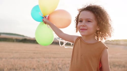 Track shot of happy boy running over countryside field and holding balloons