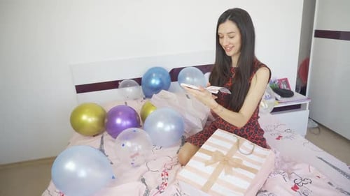 Happy Smiling Woman Holding Gift Card and Sitting on Bed with Balloons and Confetti