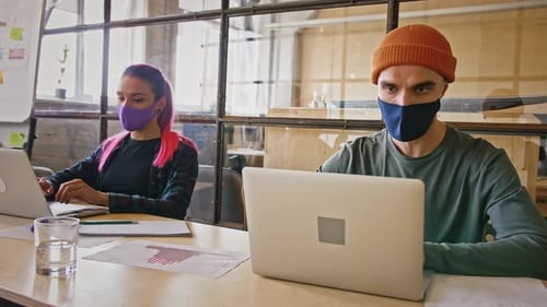 Young Coworkers Wearing Protective Medical Masks Working Oon Laptops in Office Sitting Apart of Each