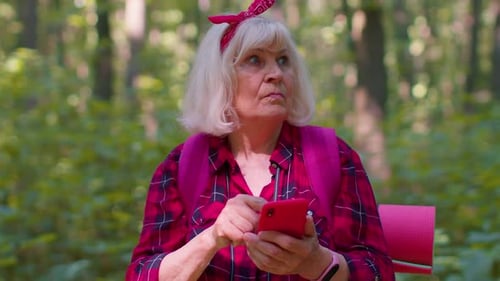 Attractive Senior Old Grandmother Woman Backpacker Planning Trip Route in Forest on Mobile Phone