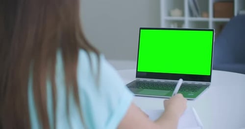 Girl Taking Notes in Notepad From Laptop Computer Green Mockup Screen. Girl Learning Online on